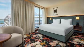 A hotel room with a large bed, a sofa, a small table, and a view of a building and water outside the window.