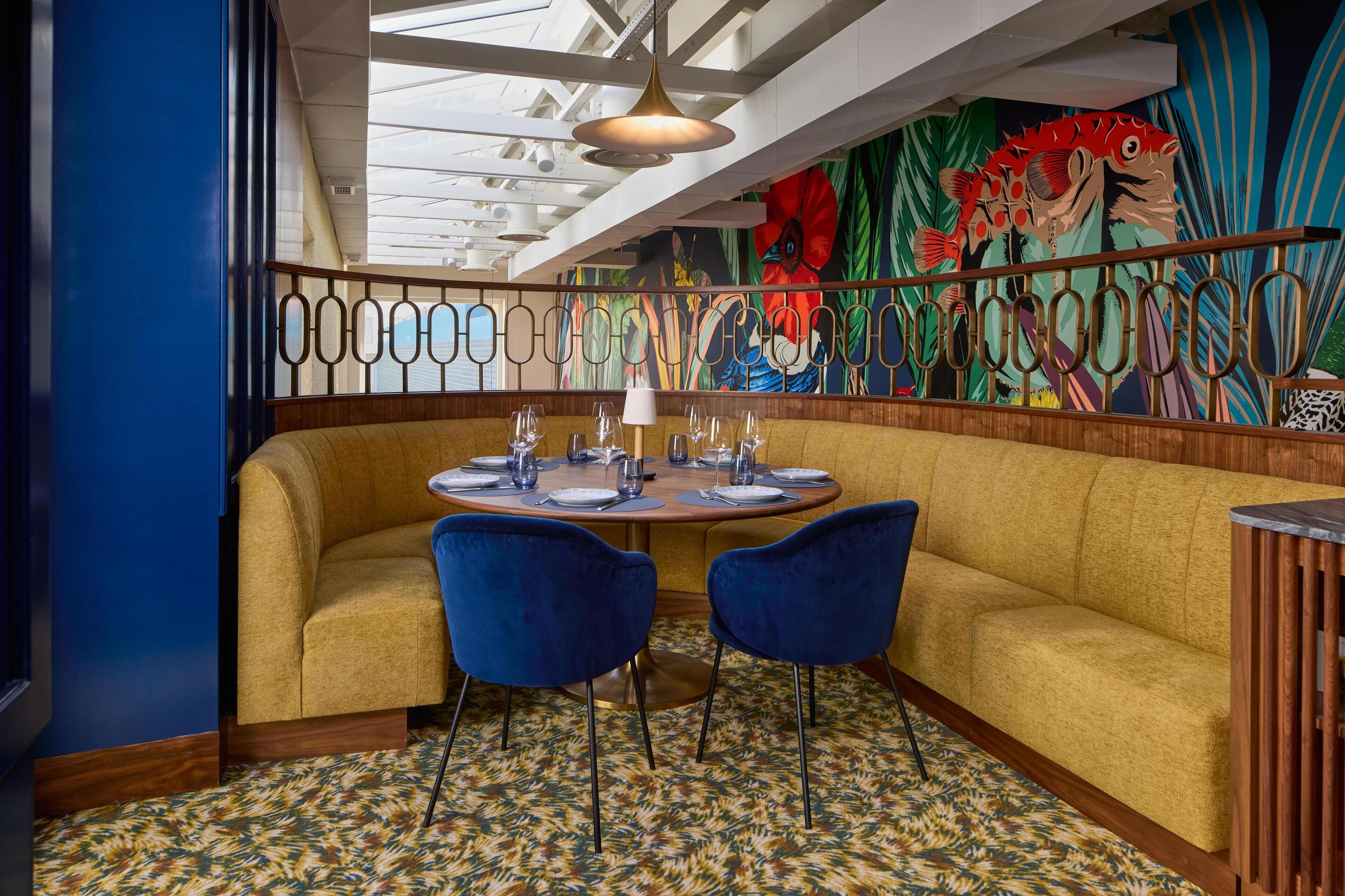 Un restaurant avec une table ronde dressée pour le repas, des chaises bleues et une fresque murale aux couleurs vives.
