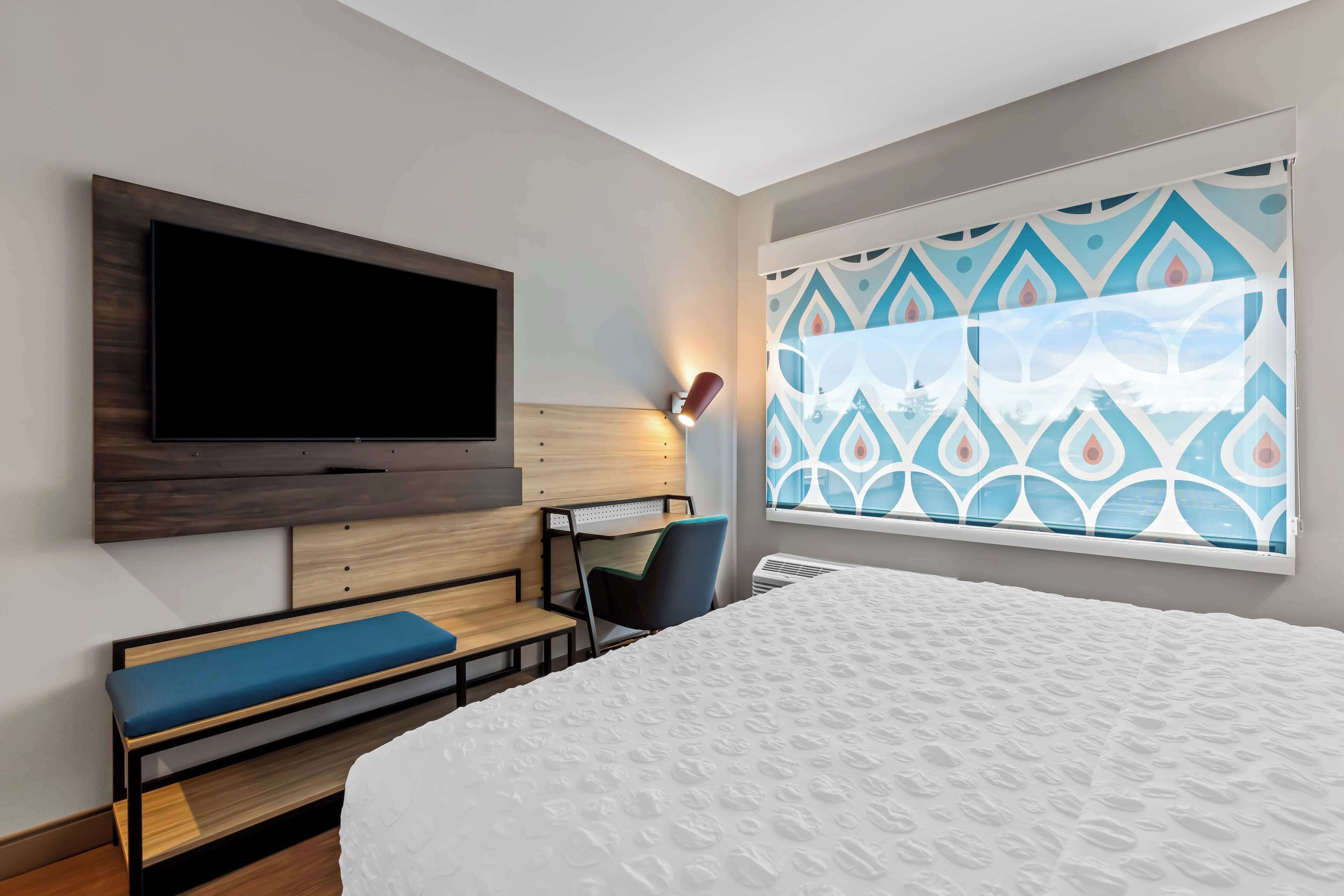 Une chambre d’hôtel dotée d’un grand lit, d’un téléviseur à écran plat fixé au mur, d’un bureau avec une chaise et d’une fenêtre agrémentée de rideaux à motifs.
