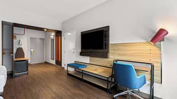 A modern hotel room with a wooden desk, a blue office chair, a flat-screen TV mounted on the wall, and a red accent lamp.