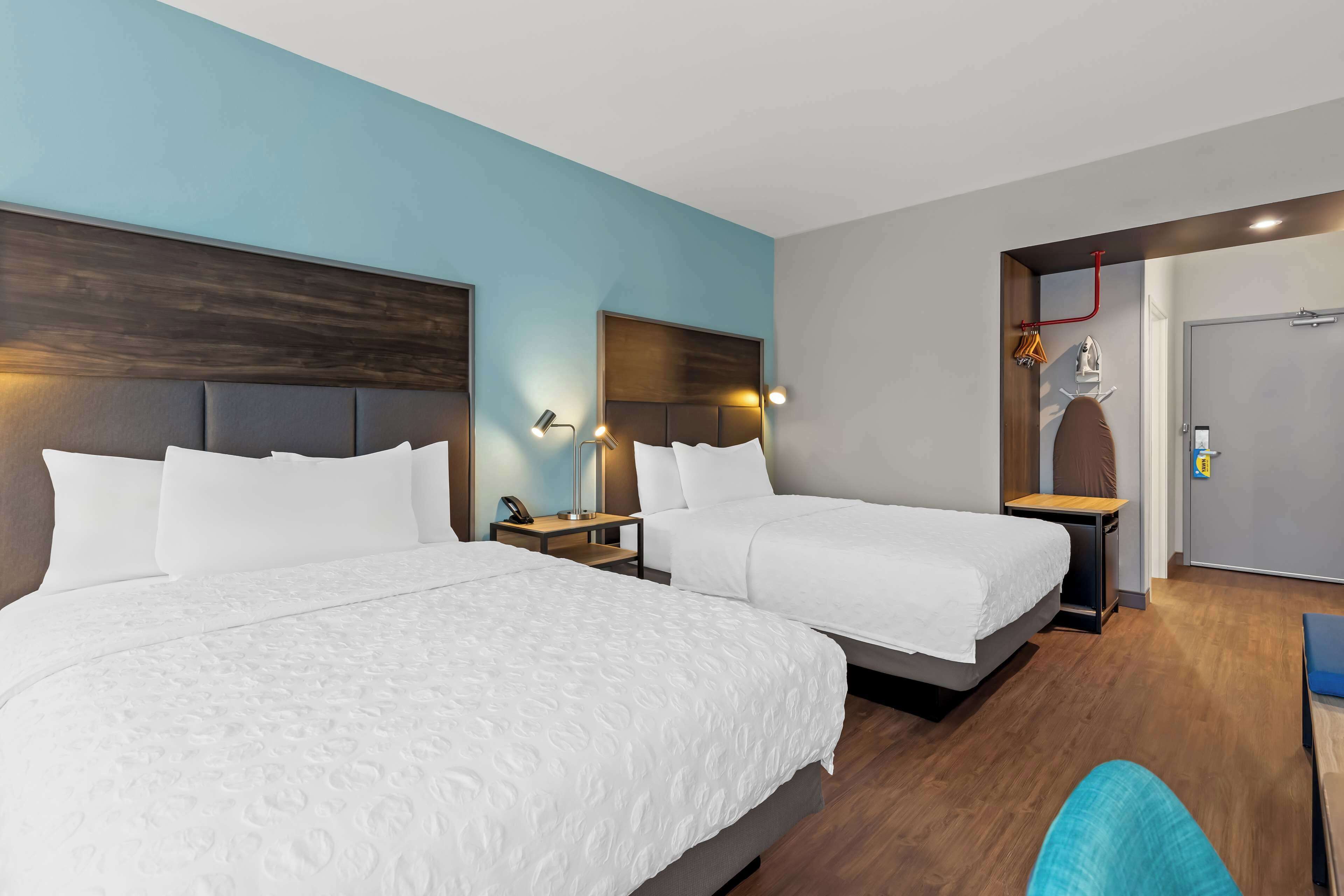 Une chambre d’hôtel avec deux lits, une tête de lit en bois, des lampes de chevet, une petite table et une porte sur laquelle est fixé un porte-serviettes.