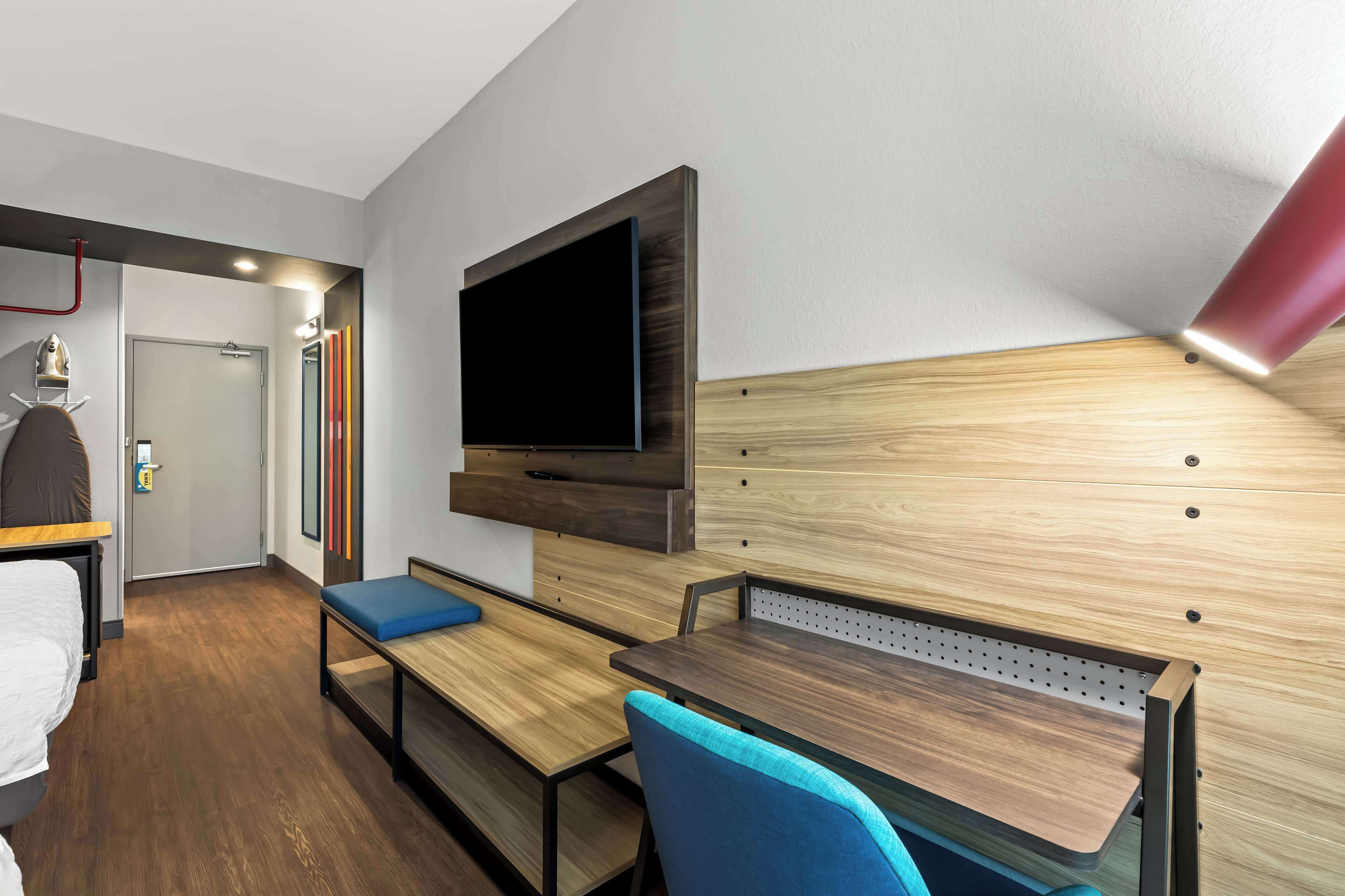 A modern hotel room with a wooden floor, a bed with a white comforter, a desk with a blue chair, a TV mounted on the wall, and a red lamp.