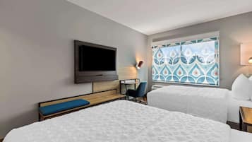 A hotel room with two beds, a TV, a desk, a chair, and a window with a patterned curtain.