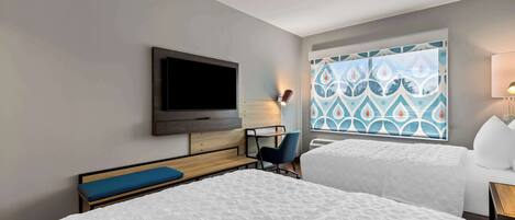 A hotel room with two beds, a TV, a desk, a chair, and a window with a patterned curtain.