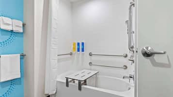 Et moderne badeværelse med et hvidt badekar, en bruser med brusehåndtag og et gelænder. Der er håndklæder hængende på en holder, og en lille hylde med toiletartikler.