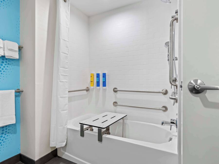 Et moderne badeværelse med et hvidt badekar, en bruser med brusehåndtag og et gelænder. Der er håndklæder hængende på en holder, og en lille hylde med toiletartikler.