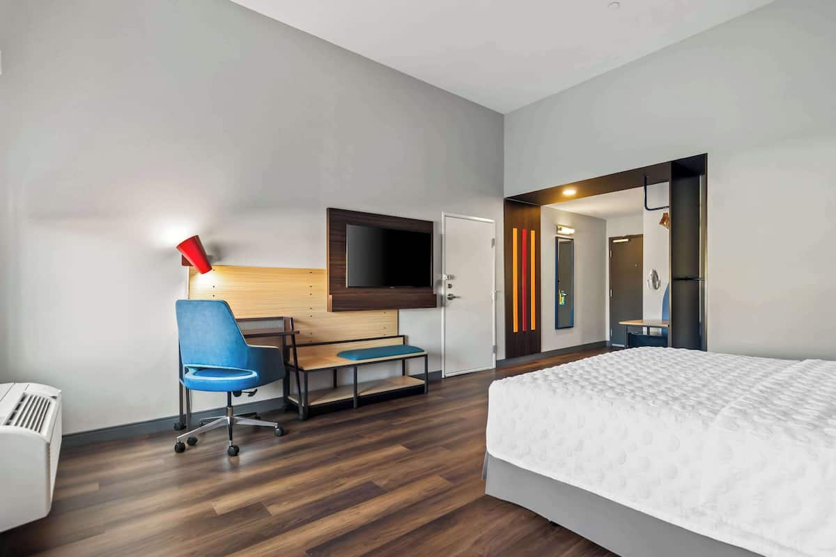 A hotel room with a bed, a desk with a chair, a TV mounted on the wall, and a door leading to another room.