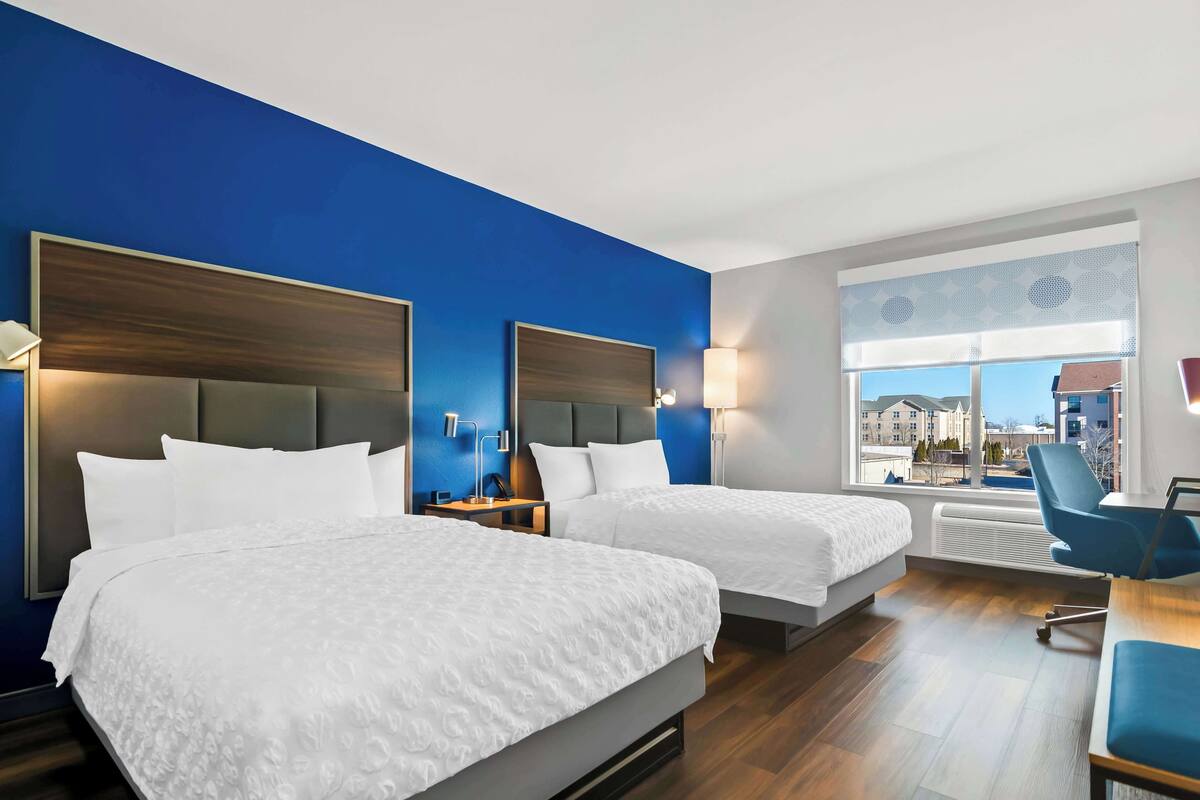A hotel room with two beds, a desk, and a chair. There is a window with a view of buildings outside.
