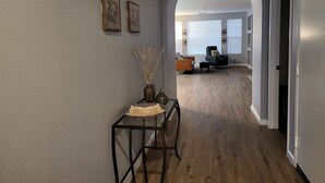 A hallway with a glass table, two framed artworks, and a door leading to a living area with a sofa and a chair. - Kid Friendly Comfy Get Away (Victorville)