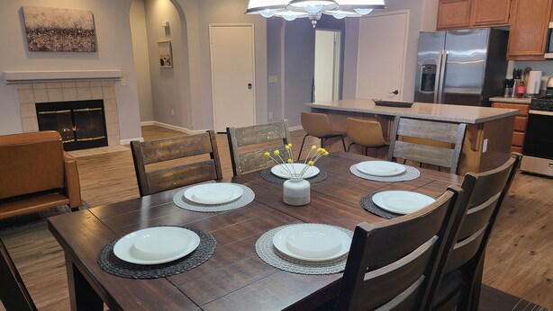 A dining area with a wooden table set for six, surrounded by chairs. A chandelier hangs above the table, and a fireplace is visible in the background. - Kid Friendly Comfy Get Away (Victorville)