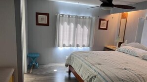 Une chambre à coucher avec un lit, un ventilateur de plafond, une fenêtre avec des rideaux et une petite table avec une lampe.