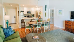 Une cuisine moderne avec des meubles blancs, une table à manger avec des chaises, un lustre et un téléviseur posé sur un meuble en bois.
