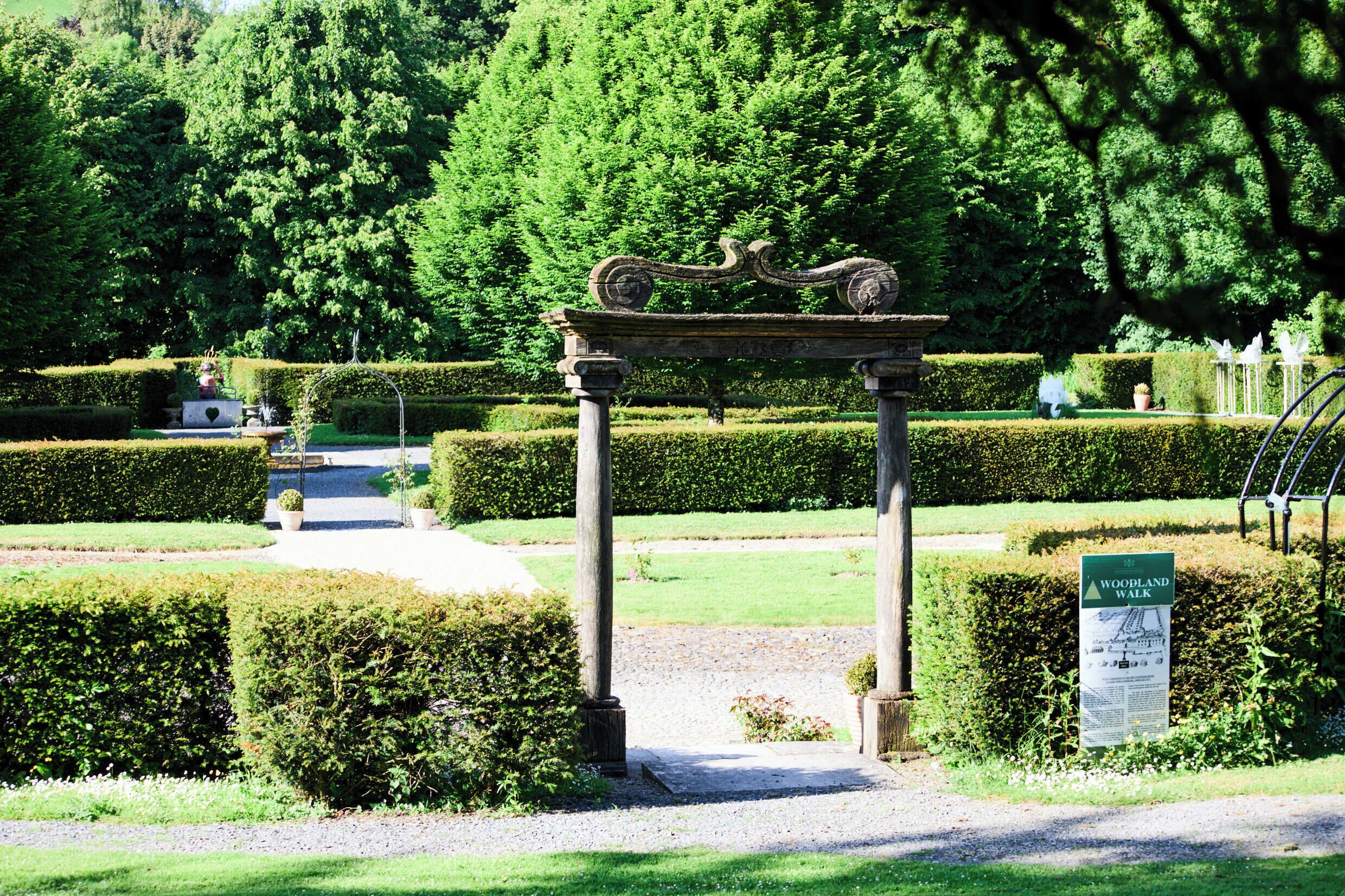 A formal garden with a central archway, neatly trimmed hedges, and a sign indicating a woodland walk.