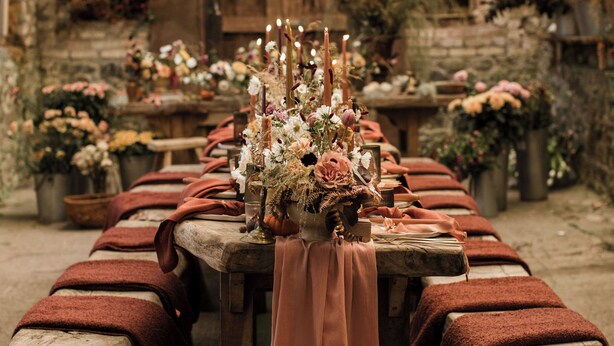 Un comedor al aire libre rústico con una larga mesa de madera decorada con flores y velas, bancos con cojines rojos y un farol colgante.
