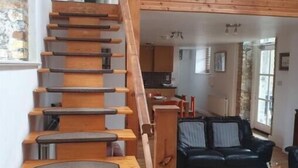 Una escalera de madera conduce a un dormitorio en buhardilla con un sofá de cuero negro y una mesa de centro de madera en la sala de estar.