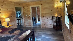 Interior de una cabaña de madera con una cama, un escritorio, una lavadora y una ventana con persianas.