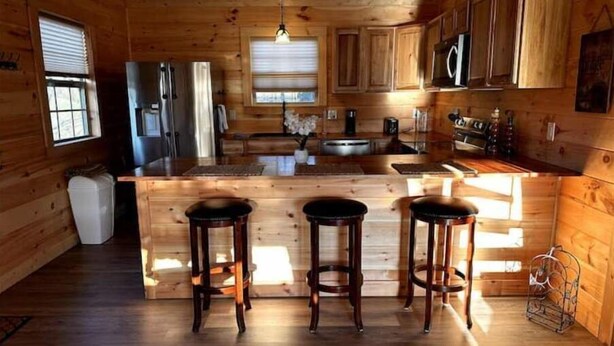 Una cabaña de madera con cocina que cuenta con una isla central con taburetes, armarios de madera y una lámpara colgante.