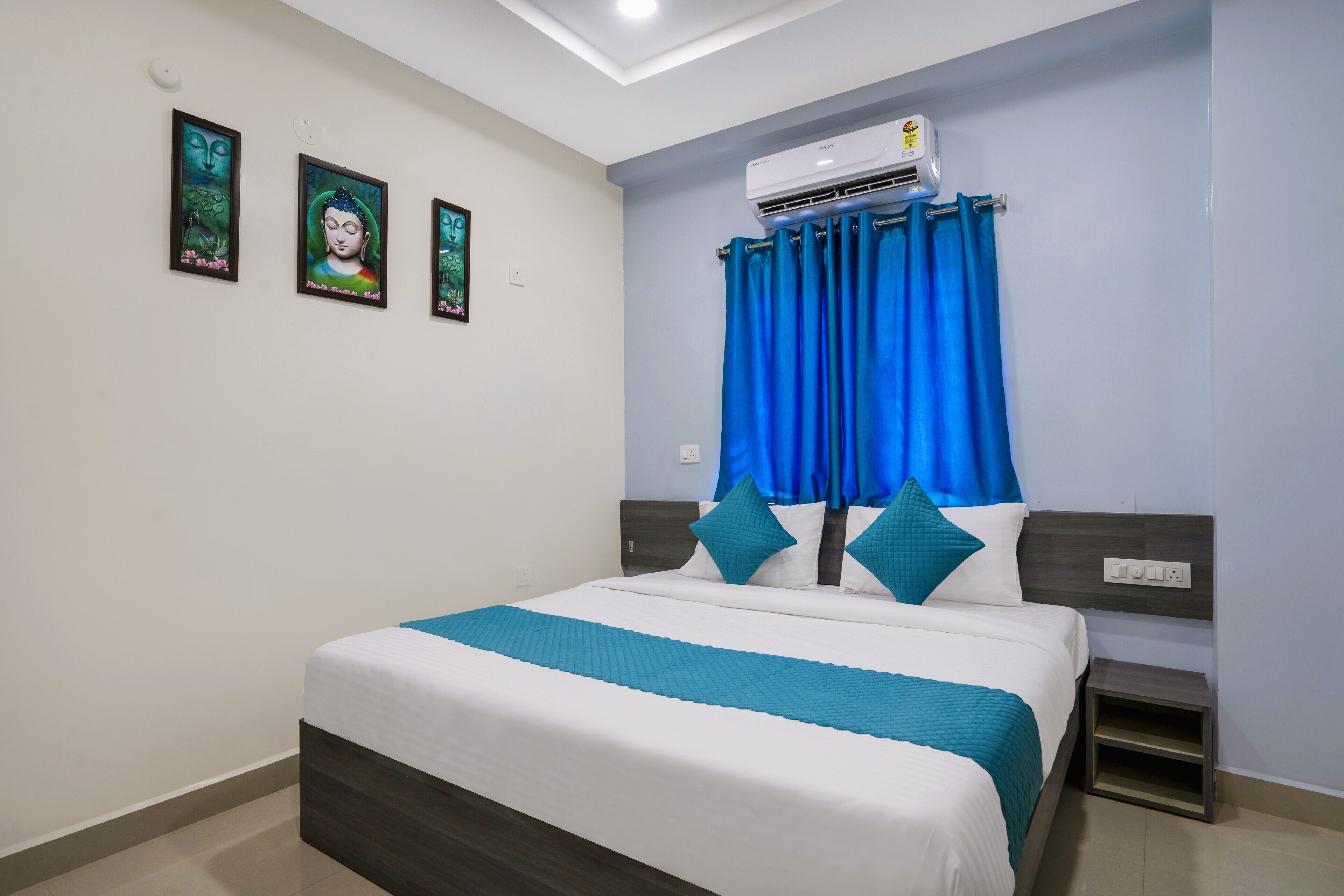 A hotel room with a bed, a nightstand, a wall-mounted air conditioner, and three framed pictures on the wall.