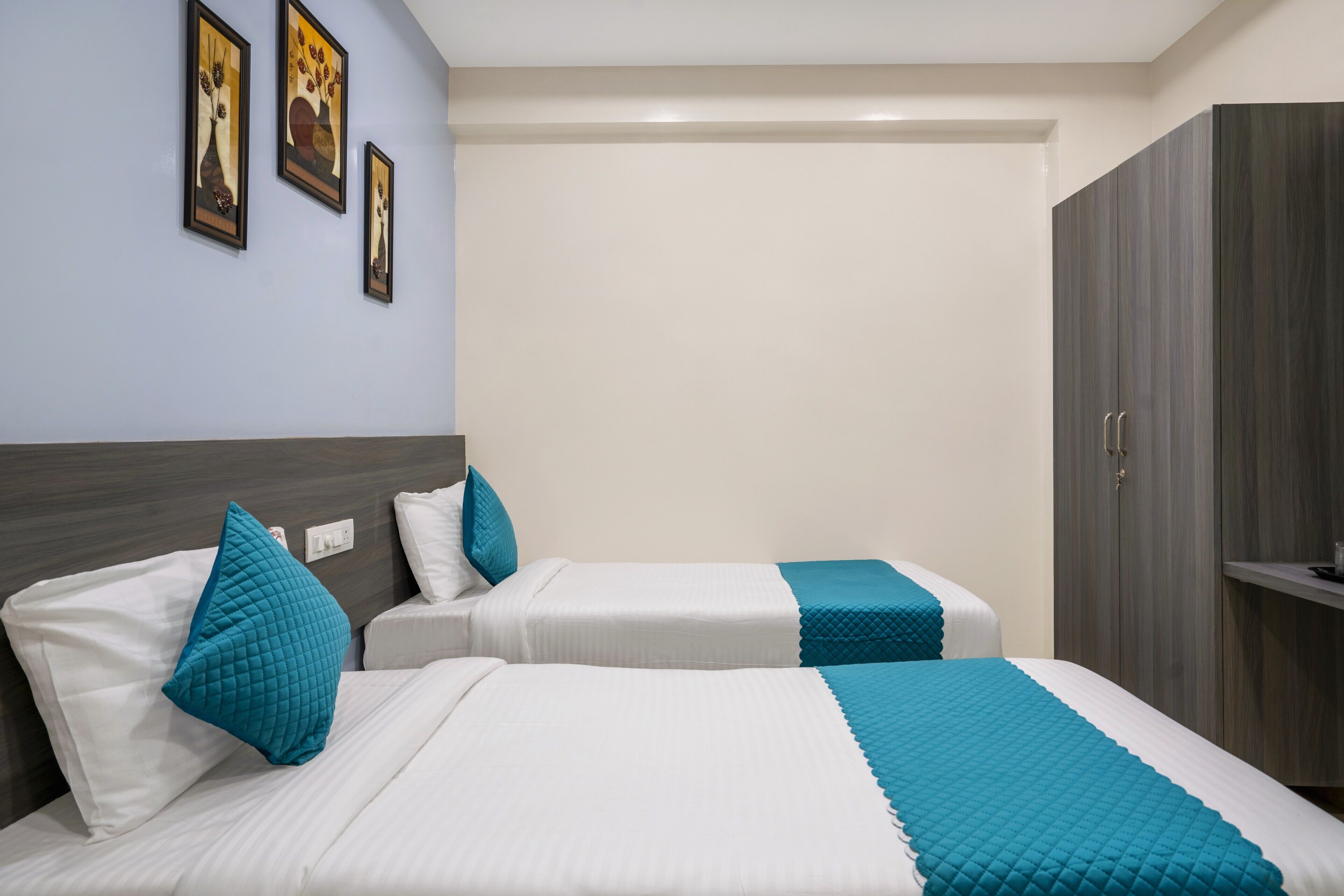 A hotel room with two beds, each with white bedding and teal accents, a wardrobe, and framed artwork on the wall.