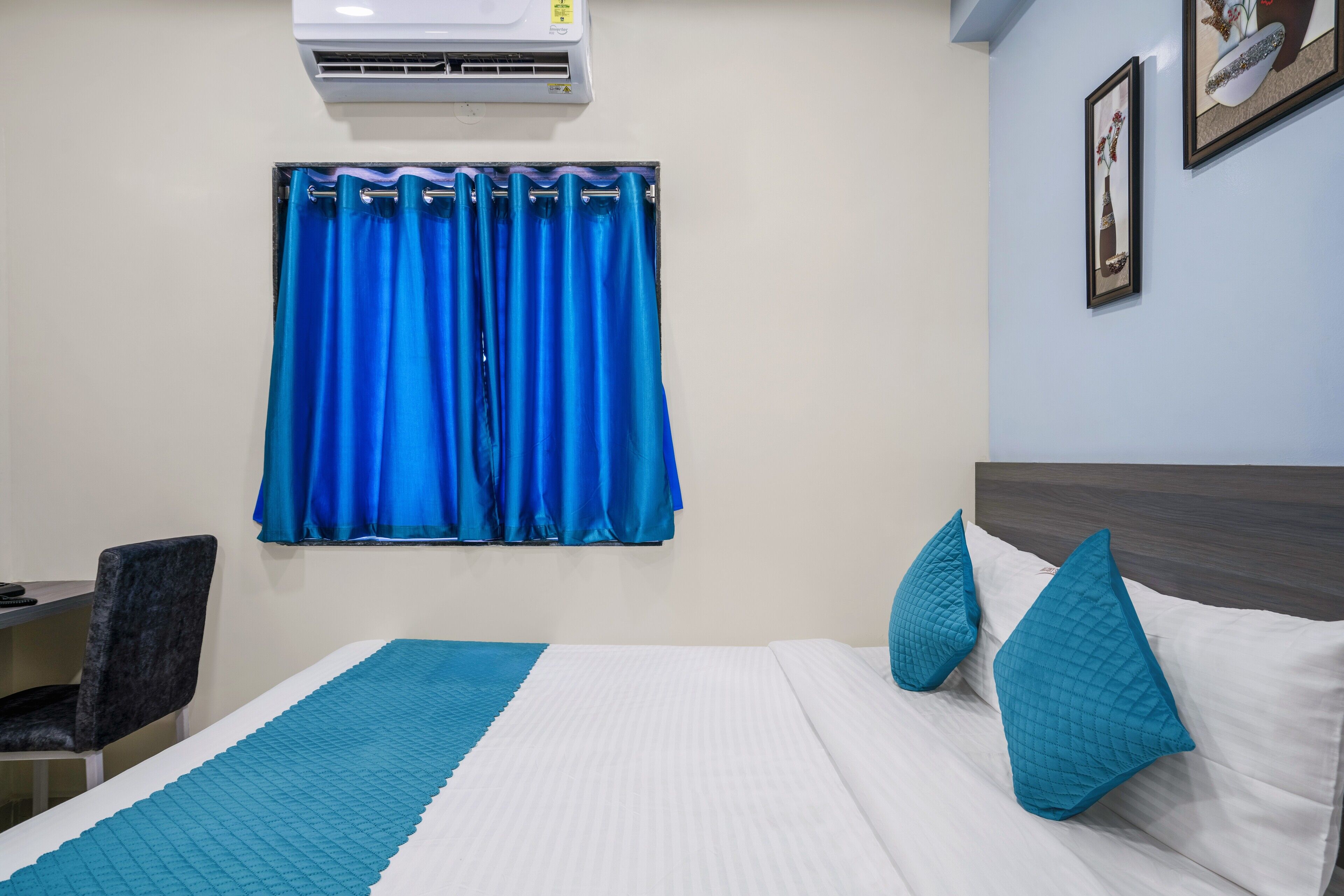 A hotel room with a bed, a desk, a chair, a window with blue curtains, and an air conditioner.