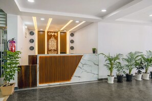 A hotel reception desk with a modern design, featuring a marble counter, wooden paneling, and multiple clocks. There are potted plants and a fire extinguisher visible. - The Balcony Suites (Hyderabad)