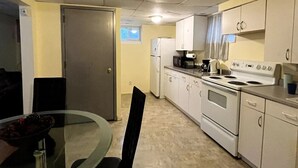 Une cuisine compacte avec des meubles blancs, un réfrigérateur, une cuisinière et un four à micro-ondes. On y trouve une table en verre avec des chaises, ainsi qu’une porte qui donne sur une autre pièce.