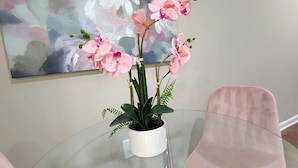A glass table with a potted orchid plant, two pink chairs, and a large abstract painting on the wall. - Cozy King Suite / Smart TVs / Self Check-In / Free Parking & Wi-Fi (Fort Mill)
