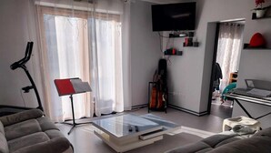 A modern living room with a glass coffee table, grey sofas, a music stand, a guitar, and a TV mounted on the wall. - Maison à Louer Août 2024, Quartier Calme, Idéal Pour les Familles (Briatexte)
