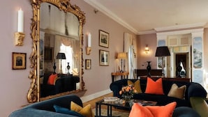Una sala de estar con un espejo grande y ornamentado, dos sofás con cojines coloridos, una mesa de centro con libros y un florero, y lámparas fijadas a la pared.