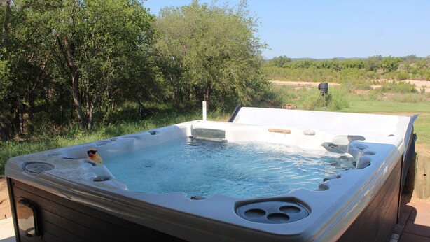 A hot tub with a wooden deck, surrounded by greenery and a clear sky.