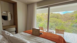 Un dormitorio con una cama grande, cabecera de madera, un espejo y vistas a zonas verdes a través de amplias ventanas.
