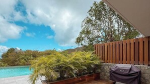 Um deck de madeira com mesa e cadeiras, churrasqueira e vista para uma piscina azul-turquesa e vegetação.