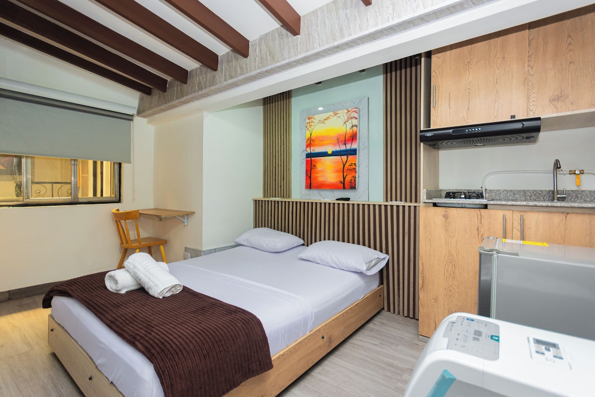 Une chambre d’hôtel avec un lit, une tête de lit en bois, un tableau, un coin cuisine et une chaise.