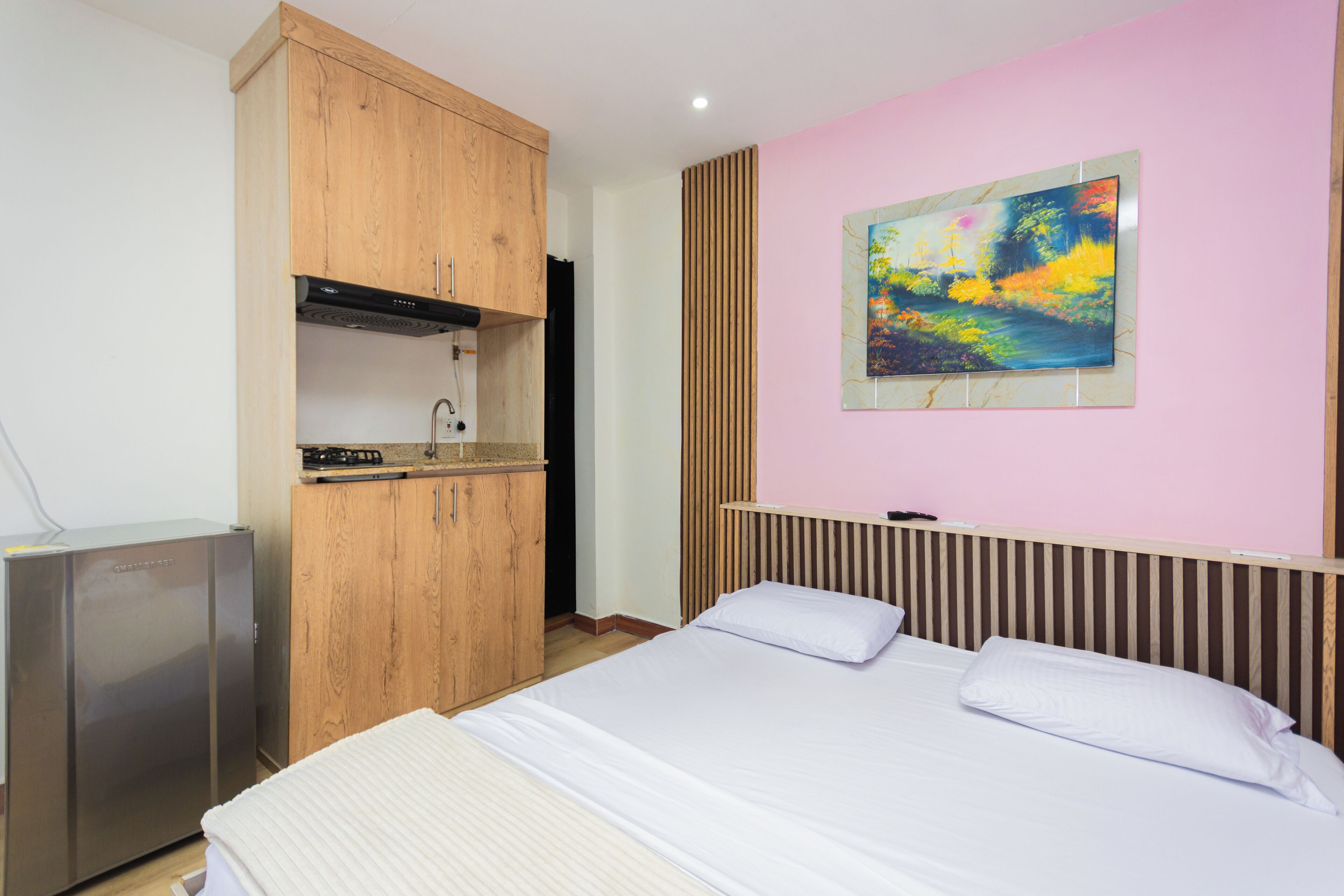 A bedroom with a bed, a wooden wardrobe, a built-in kitchen area, and a painting on the wall.