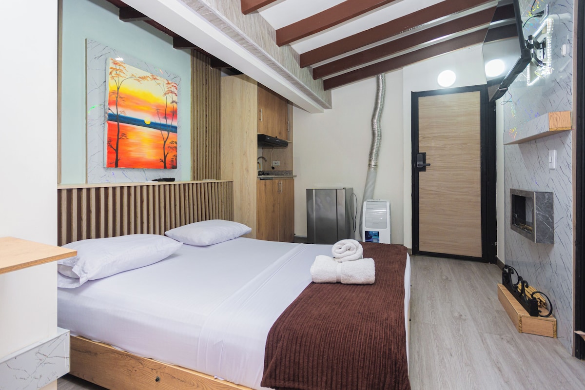 A hotel room with a bed, a painting on the wall, a wooden headboard, a wooden bench, a towel, a heater, and a door.