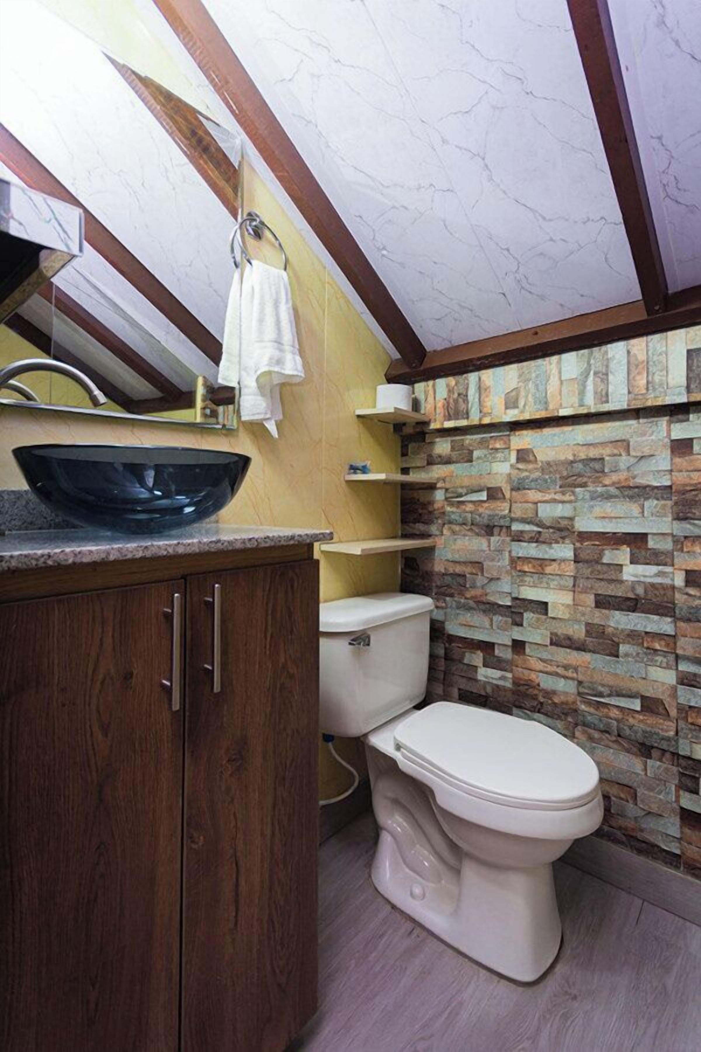 Une salle de bain avec un mur recouvert de pierres derrière le lavabo, un meuble-lavabo en bois, un lavabo sur pied noir et des toilettes.