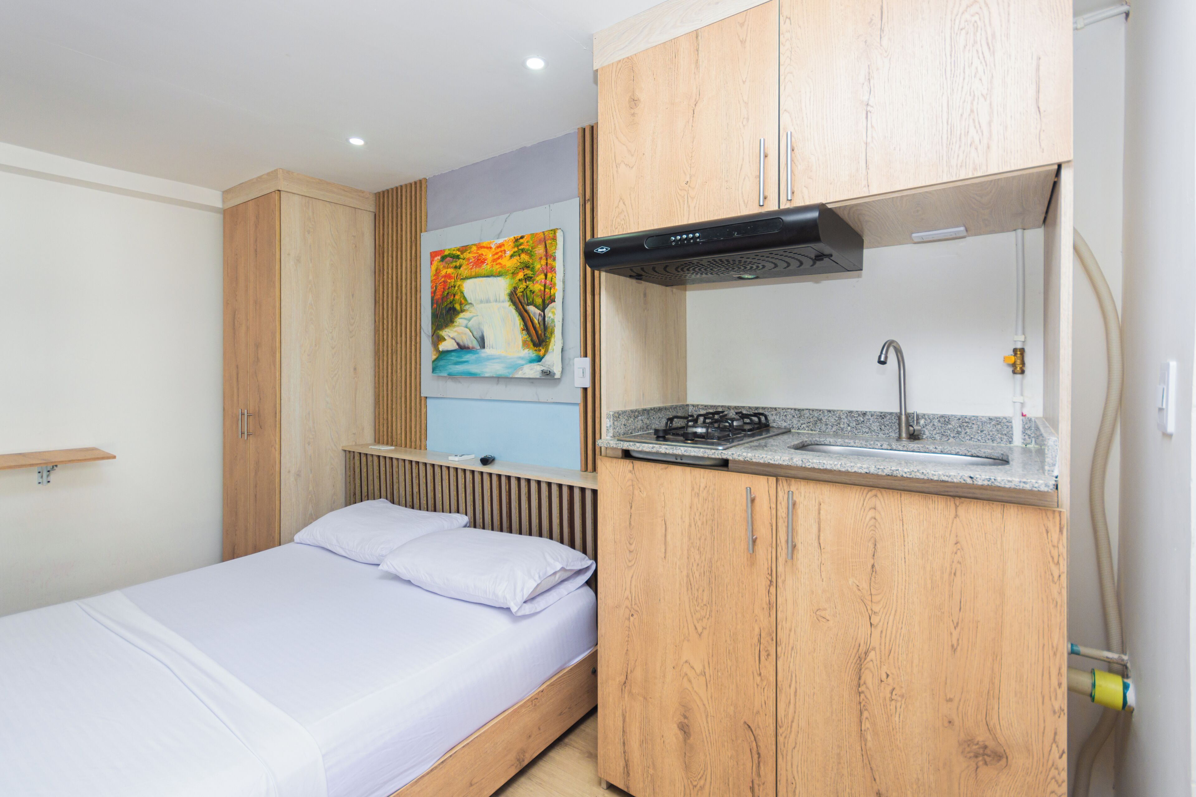 A compact room with a bed, wooden cabinets, a small kitchenette with a gas stove, and a painting on the wall.