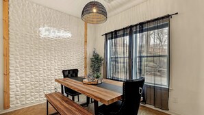 Un coin repas avec une table en bois, des chaises noires, un banc, un luminaire suspendu et une fenêtre ornée de rideaux légers.