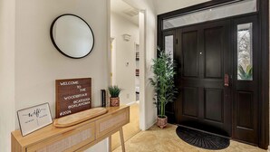 Un couloir avec une porte en bois sombre, un miroir rond, une pancarte de bienvenue, une console sur laquelle est posé un routeur, et une plante en pot.