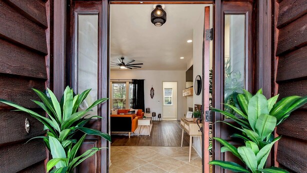 Une façade en bois avec une porte vitrée donnant sur un intérieur comprenant un coin repas, un ventilateur de plafond et des plantes en pot.