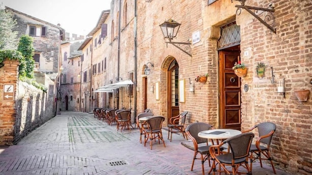 A narrow cobblestone street lined with traditional brick buildings, outdoor seating with wicker chairs and tables, and hanging lanterns.