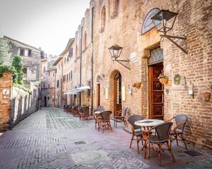 A narrow cobblestone street lined with traditional brick buildings, outdoor seating with wicker chairs and tables, and hanging lanterns. - Hotel Il Castello (Certaldo)
