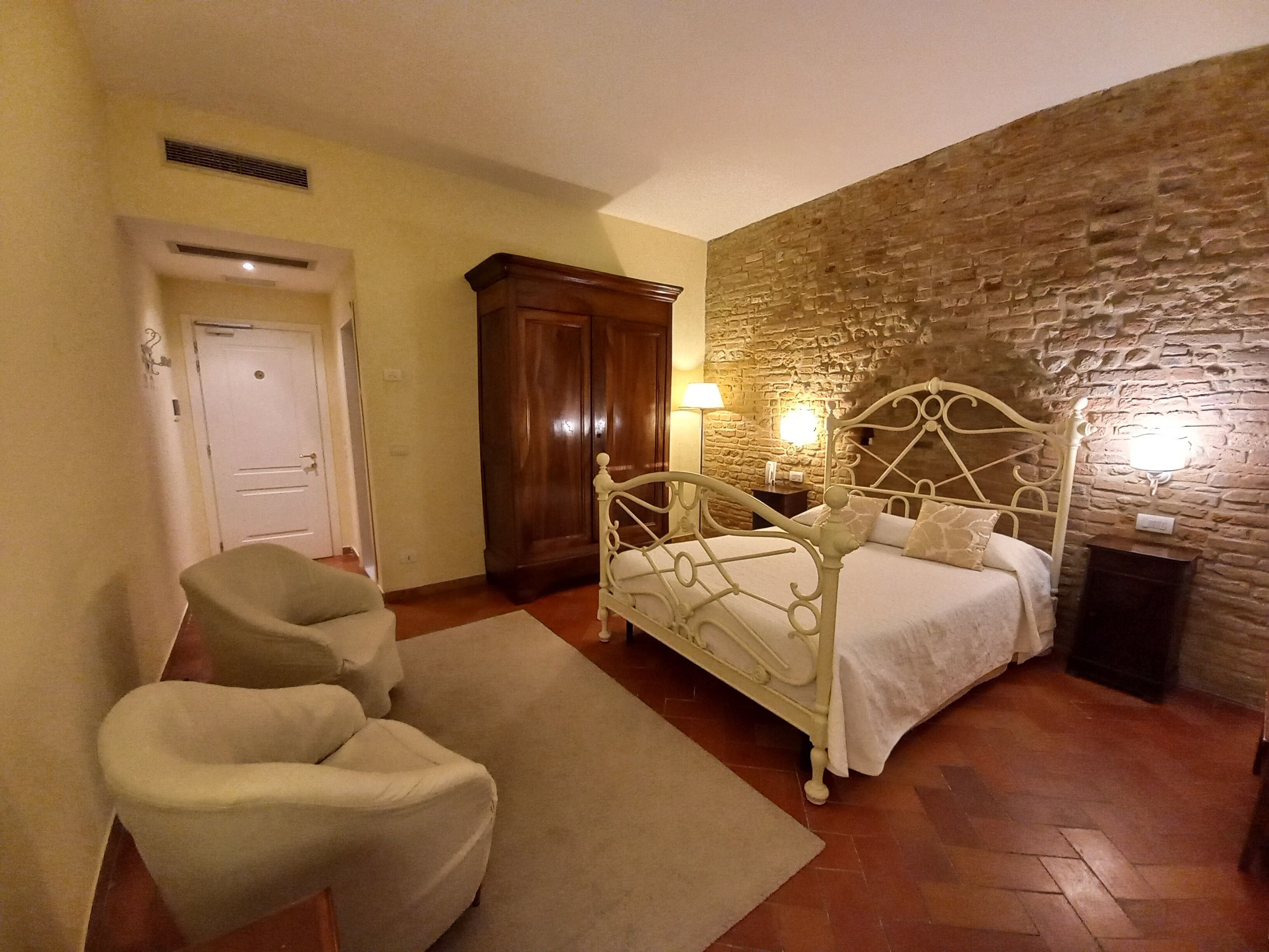 A bedroom with a stone wall, a bed with a headboard, a chair, and a nightstand with a lamp.