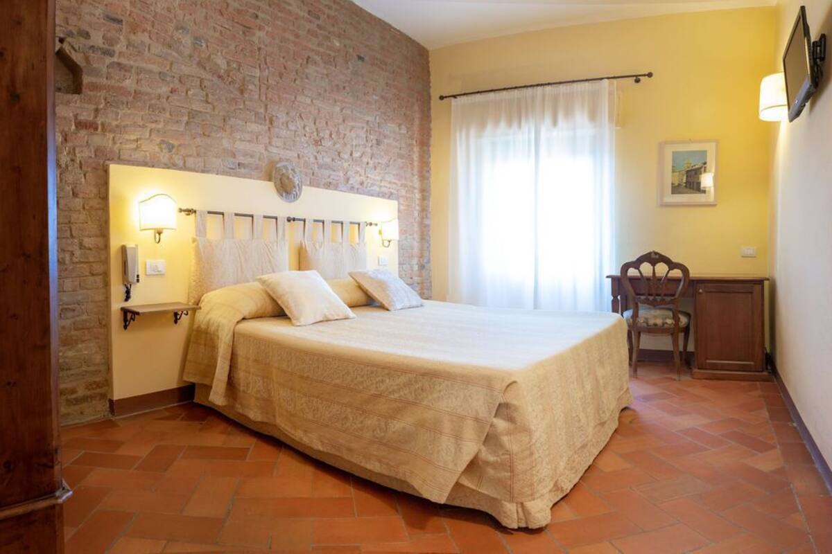 Un dormitorio con pared de ladrillo, una cama con cabecera, una mesita de noche, una silla y una ventana con cortinas.
