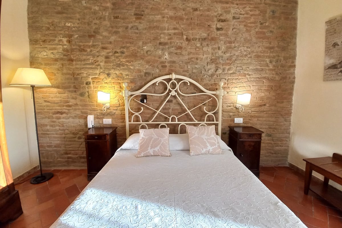 Un dormitorio con pared de ladrillo, estructura de cama metálica, dos mesitas de noche y una lámpara de pie.
