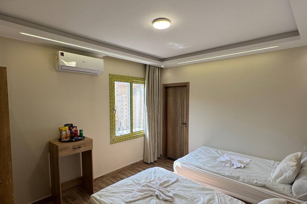 A room with two beds, a small table with snacks, an air conditioning unit, a window with curtains, and a door.