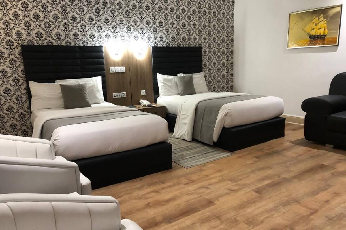 A hotel room with two beds, a sofa, a chair, a wooden floor, and a patterned wallpaper.