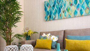 Een woonkamer met een bank, gele en blauwe kussens, een grote plant en decoratieve vazen op een houten salontafel.