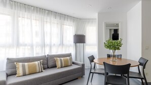 Un salon moderne avec un canapé gris, deux coussins, une table ronde en bois accompagnée de quatre chaises, un grand lampadaire et une plante en pot.