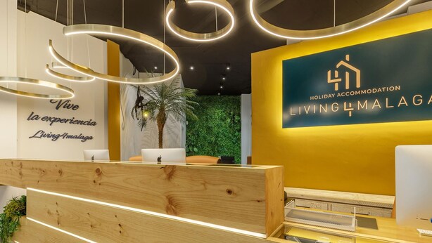 A reception area with a modern design, featuring a wooden reception desk, a green wall with a palm tree, and a sign that reads "Holiday Accommodation Living4Malaga".